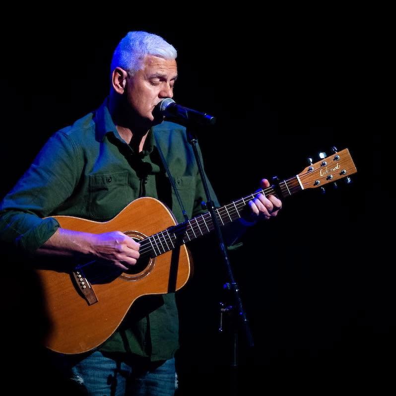 Ryan Edwards singing into a microphone with an acoustic guitar on a darkened stage